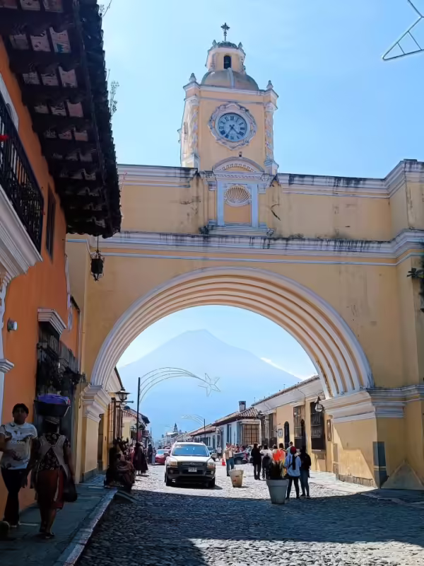 Daytime street scene featuring Arco de Santa Catalina (Saint Catherine's Arch) in Antigua Guatemala, with Volcán de Fuego seen through the arch